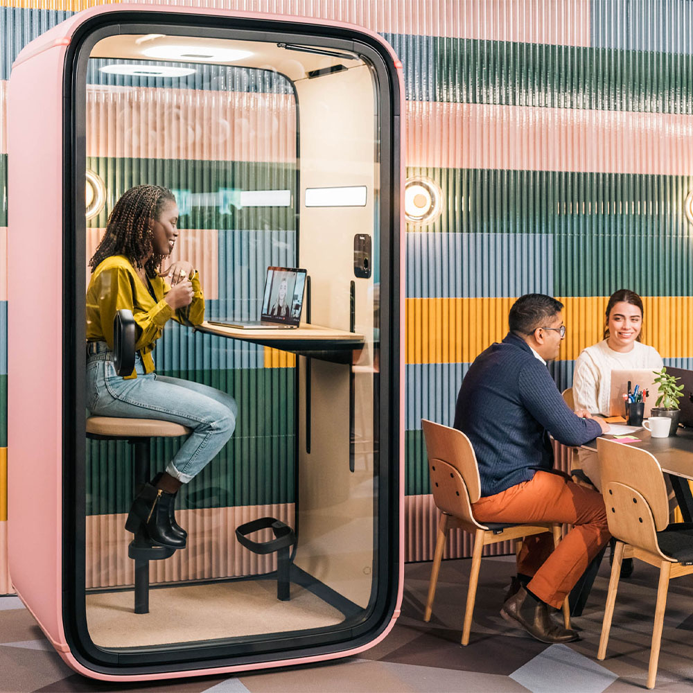 A person in a video conference in a Framery One office pod while other people are sitting at a table next to the pod.