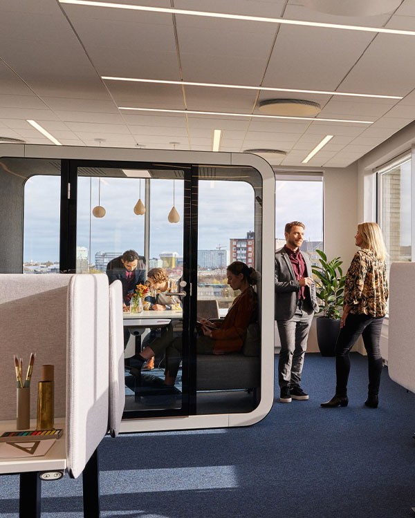 Office workers having a conversation next to a Framery Q meeting pod, in which people are working.
