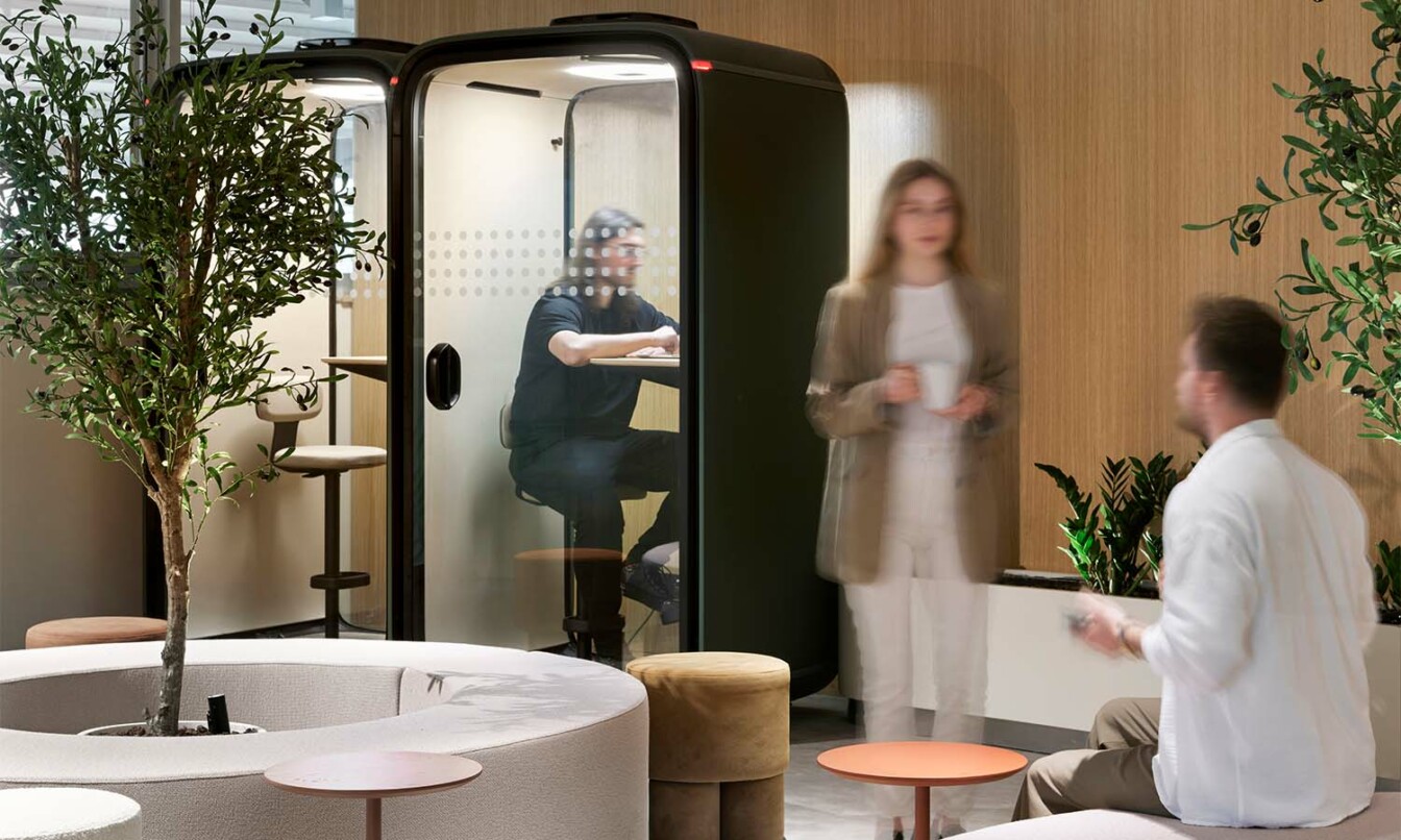 Framery One soundproof office pod in an open space with a person working inside of it while others have a conversation just next to it.